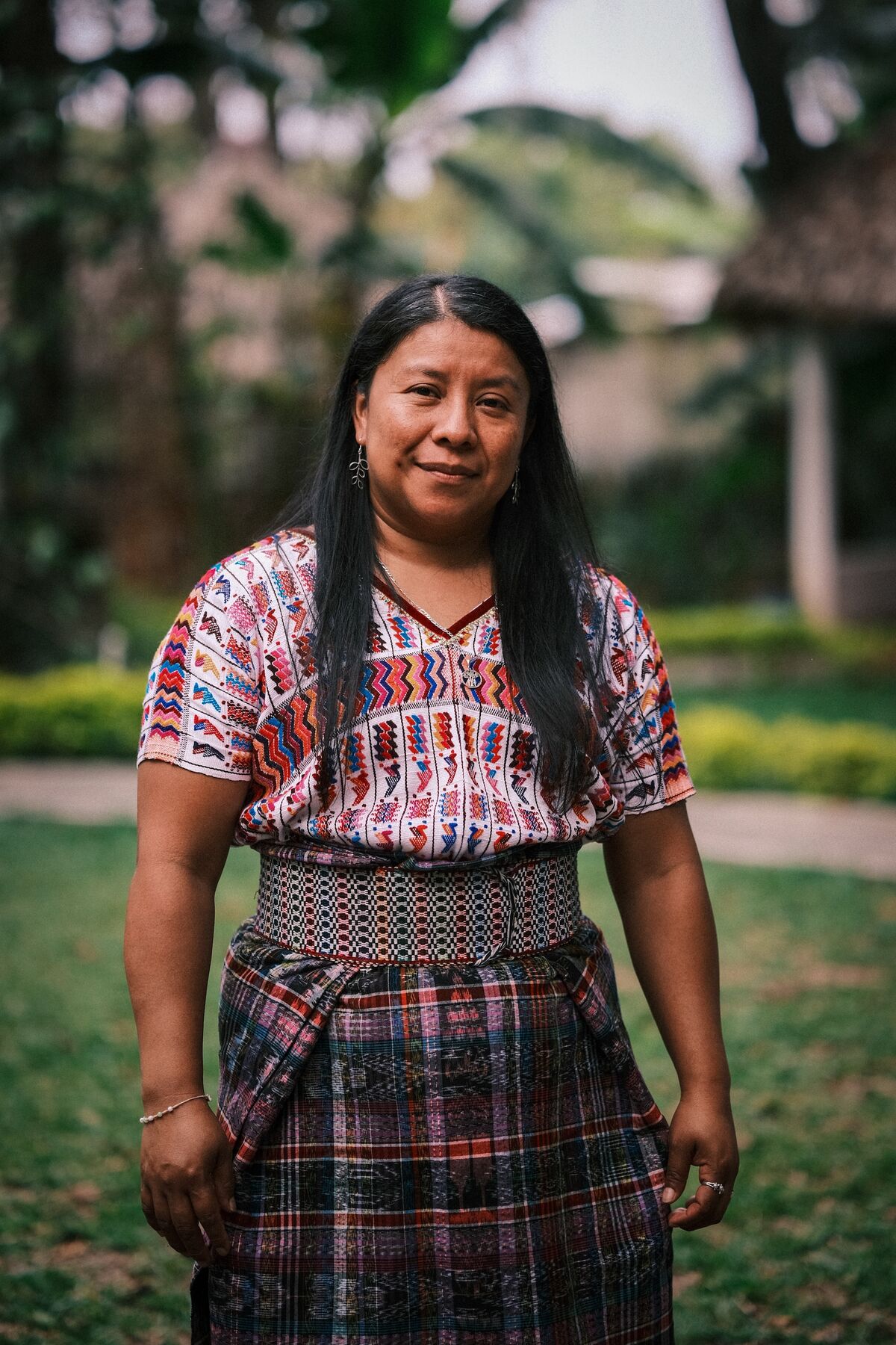 A woman in a traditional embroidered huipil and woven skirt, standing in a garden