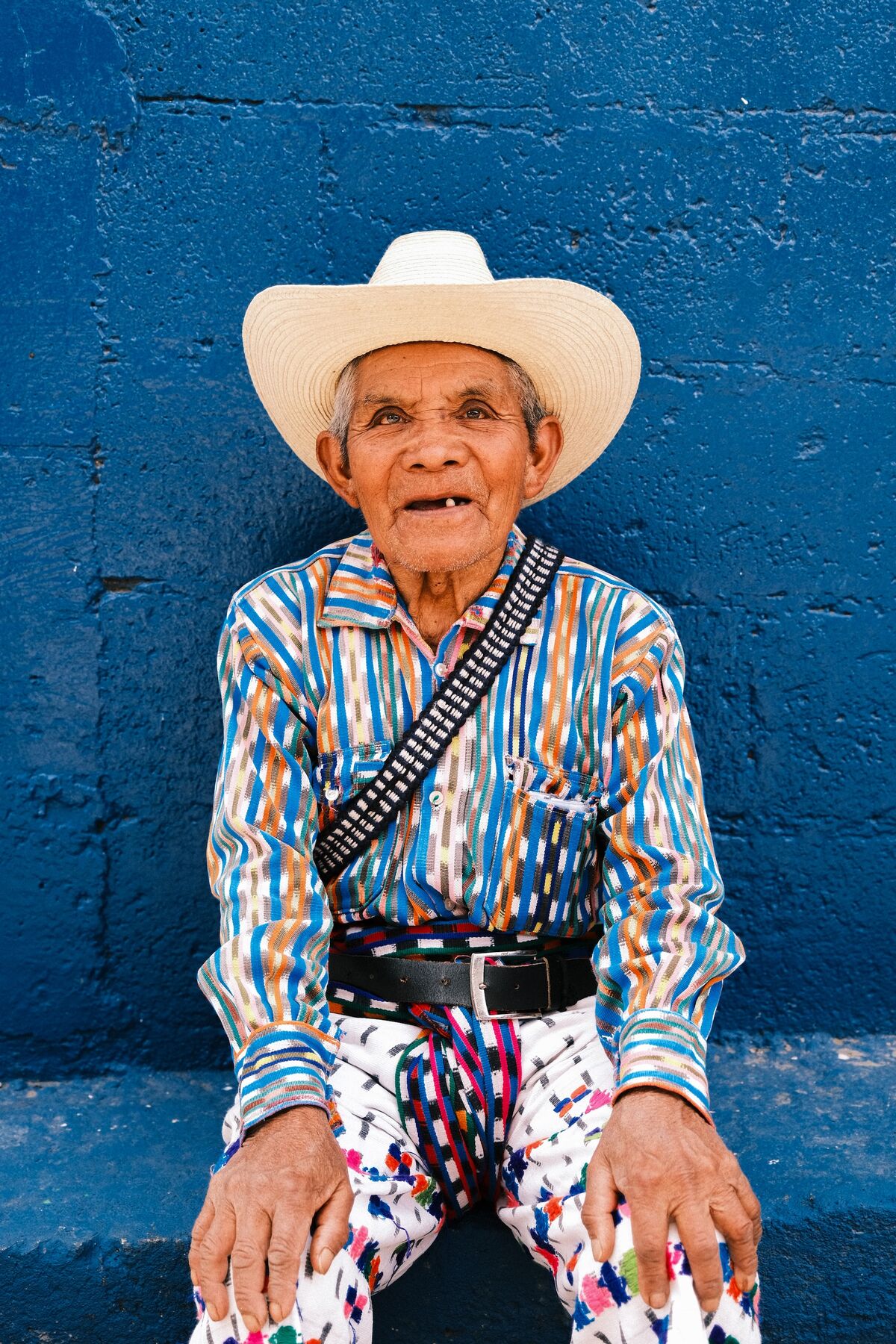 An older man in a straw hat and striped traditional shirt, seated in front of a blue wall