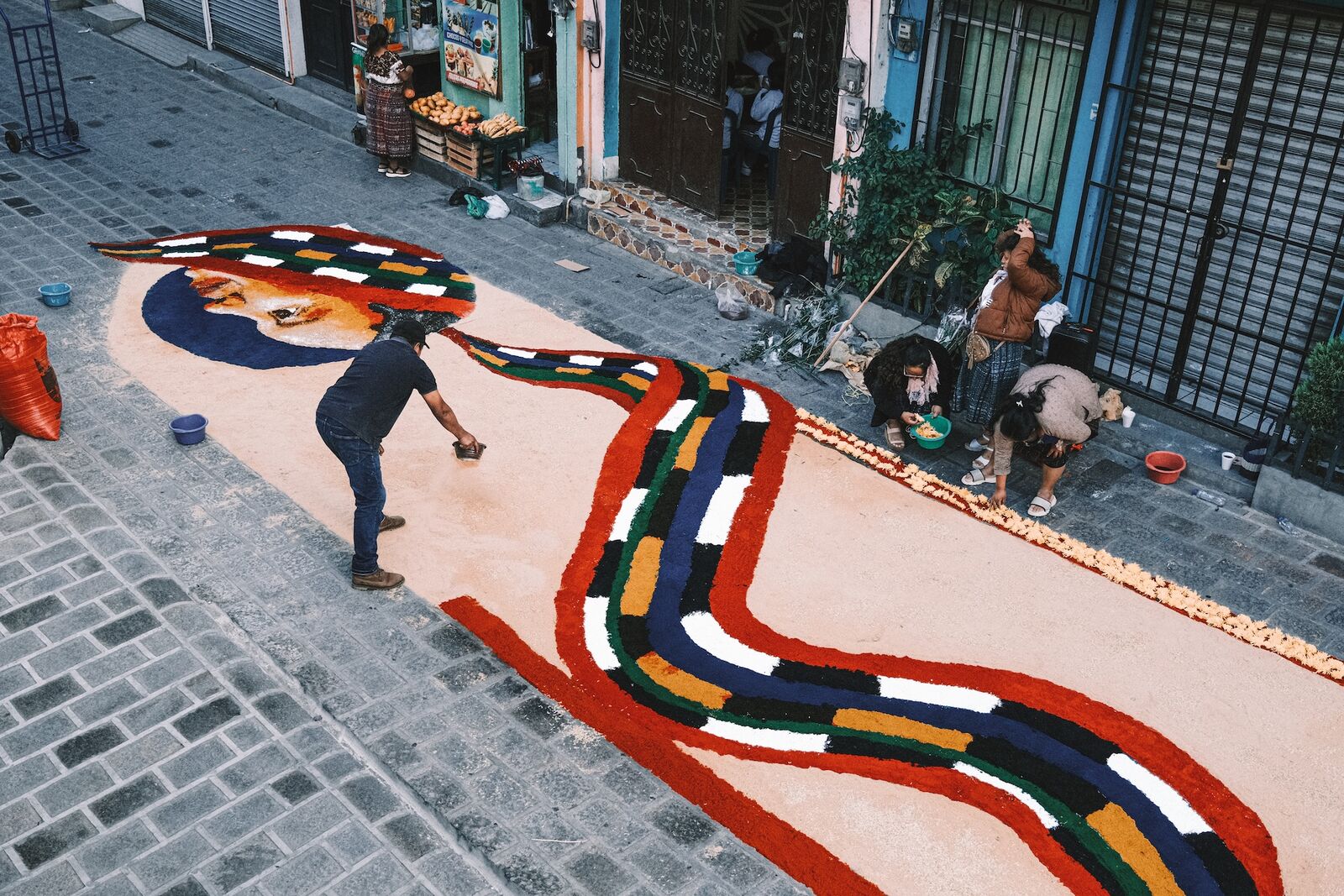 A man laying a colorful sawdust carpet with a face and striped bands across a cobbled street while neighbors prepare flowers