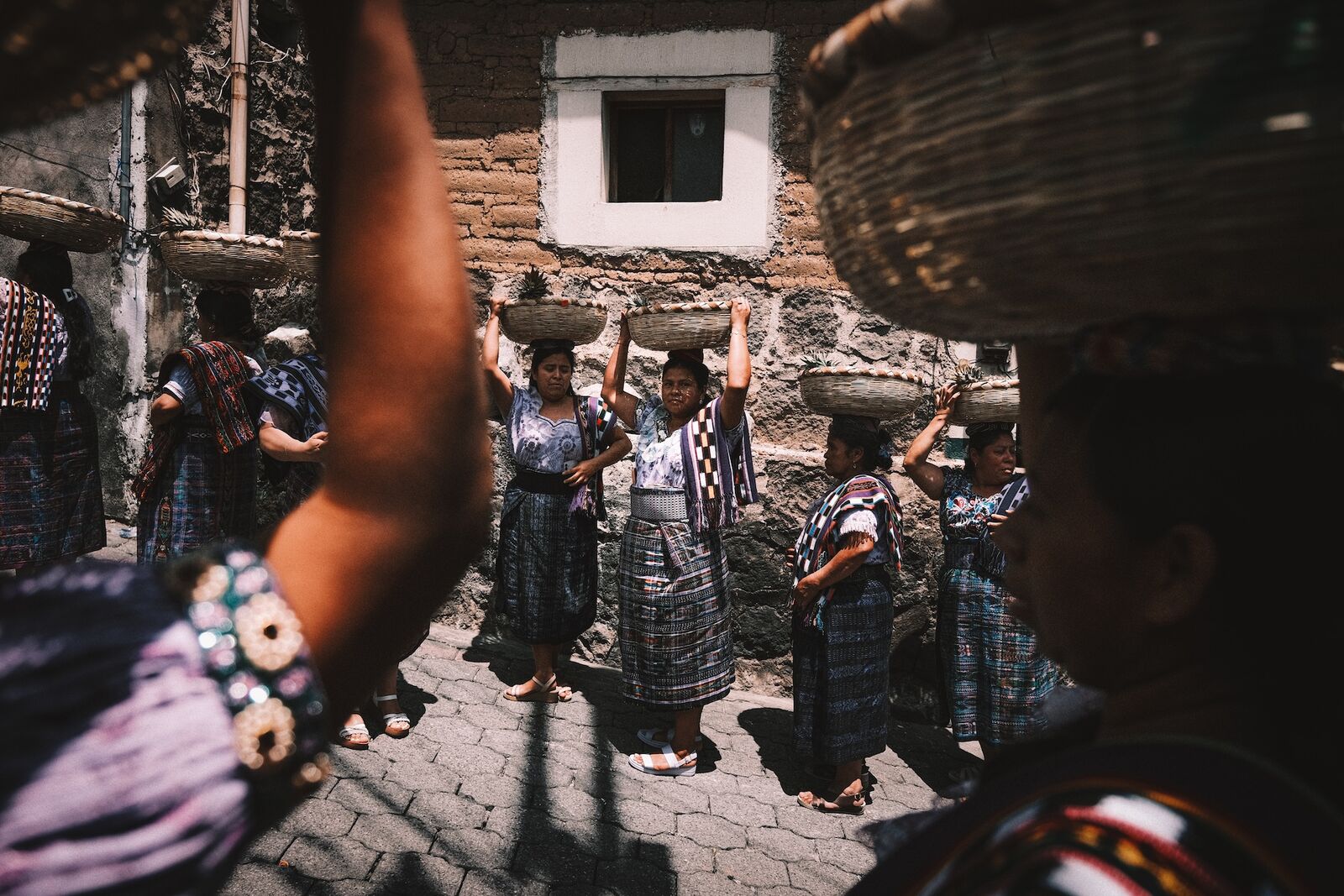 Women in traditional dress carrying baskets of fruit on their heads through a narrow street during a procession