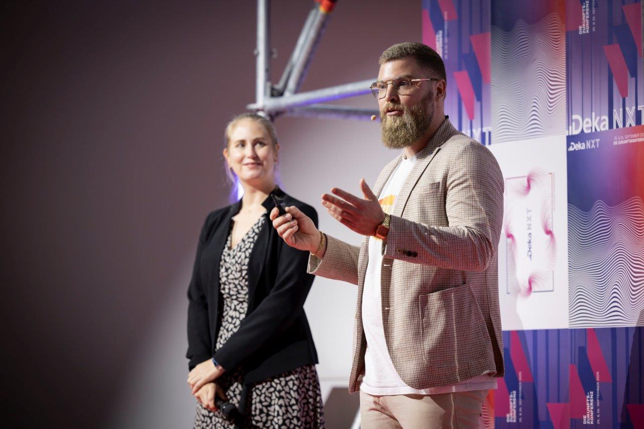 Timo Strohmann speaking on stage at DekaNXT 2025, with Doreen Mahnke beside him